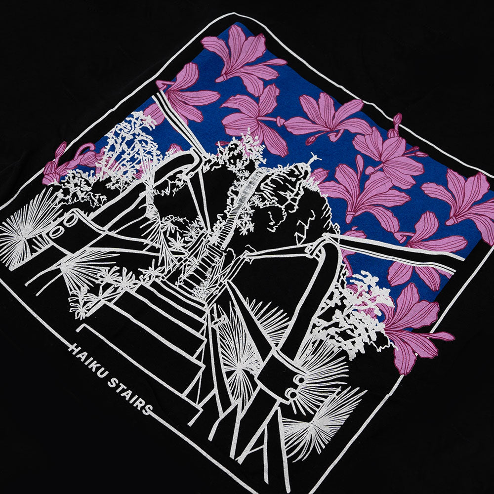 Close-up of the back design on a black t-shirt featuring a white line illustration of the Haʻikū Stairs (also known as the Stairway to Heaven) in Hawaii, surrounded by tropical plants. The background is filled with vibrant pink flowers and a blue sky within a rectangular frame. The words "HAIKU STAIRS" appear at the bottom of the design.