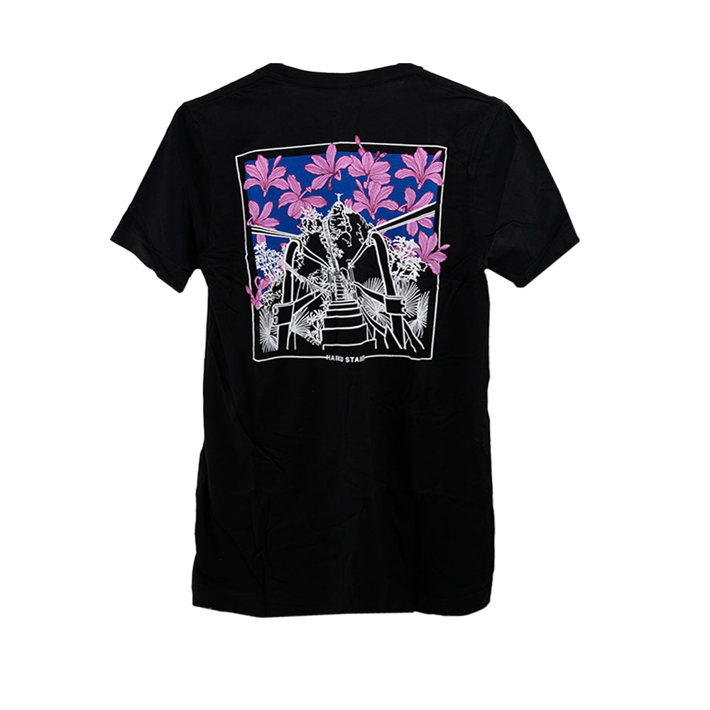 Black t-shirt featuring a vibrant back graphic of the Haiku Stairs illustrated in white lines. The stairs are surrounded by tropical foliage, with bold pink and purple flowers set against a gradient blue sky background. The text "HAIKU STAIRS" is subtly printed near the bottom of the design.