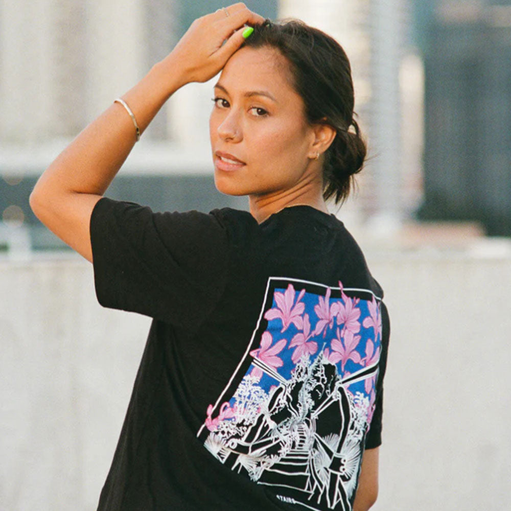 A person wearing a black t-shirt with a colorful back design featuring the Haiku Stairs outlined in white. The stairs are surrounded by pink and purple tropical flowers against a blue gradient sky. The person has dark hair pulled back, is looking over their shoulder, and has one hand raised to their head, posing in an outdoor urban setting.