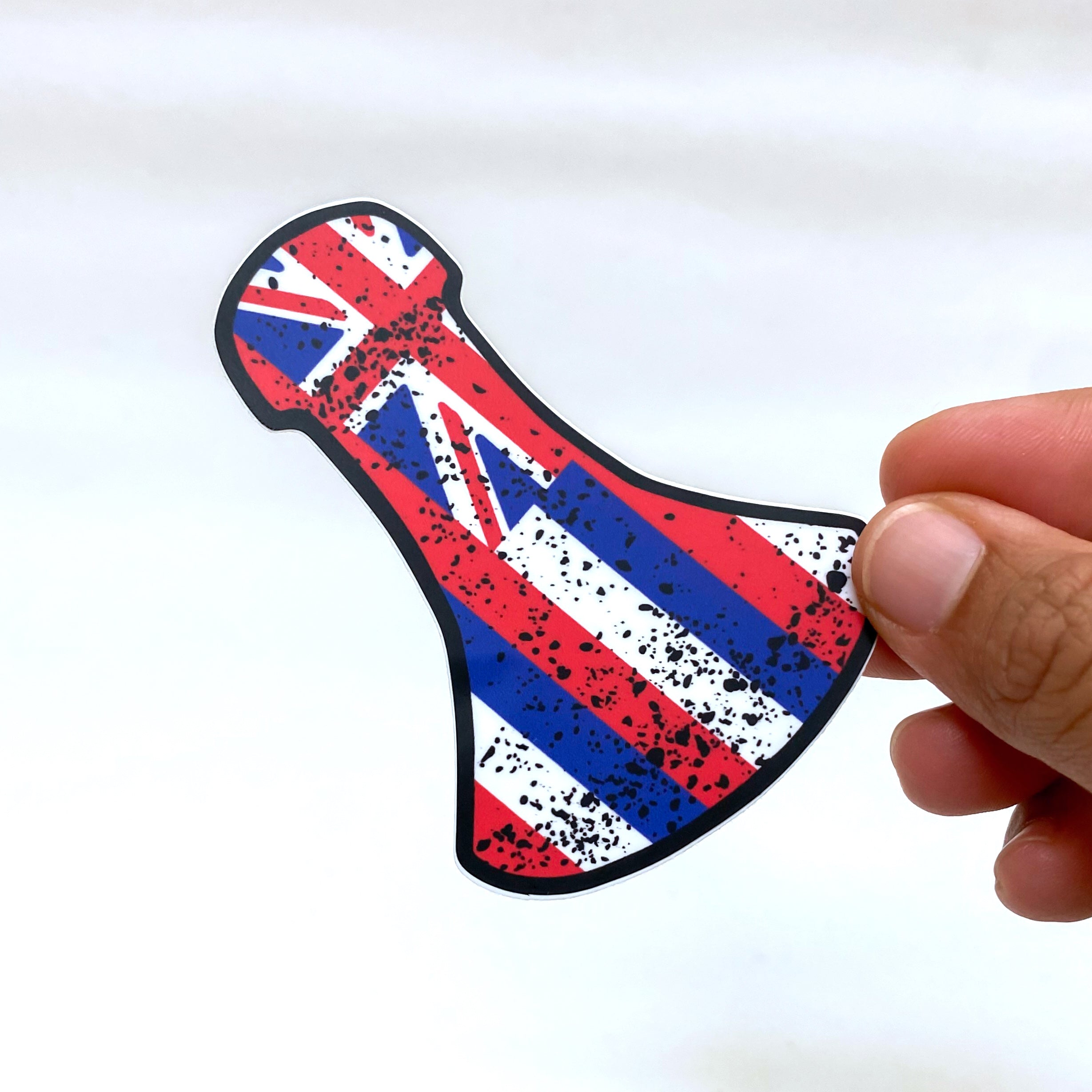 A hand holds a sticker shaped like a Hawaiian war club (lei o mano), decorated with the Hawaiian flag design in red, white, and blue. The surface features a distressed, splattered black texture for a weathered appearance. The background is plain white.