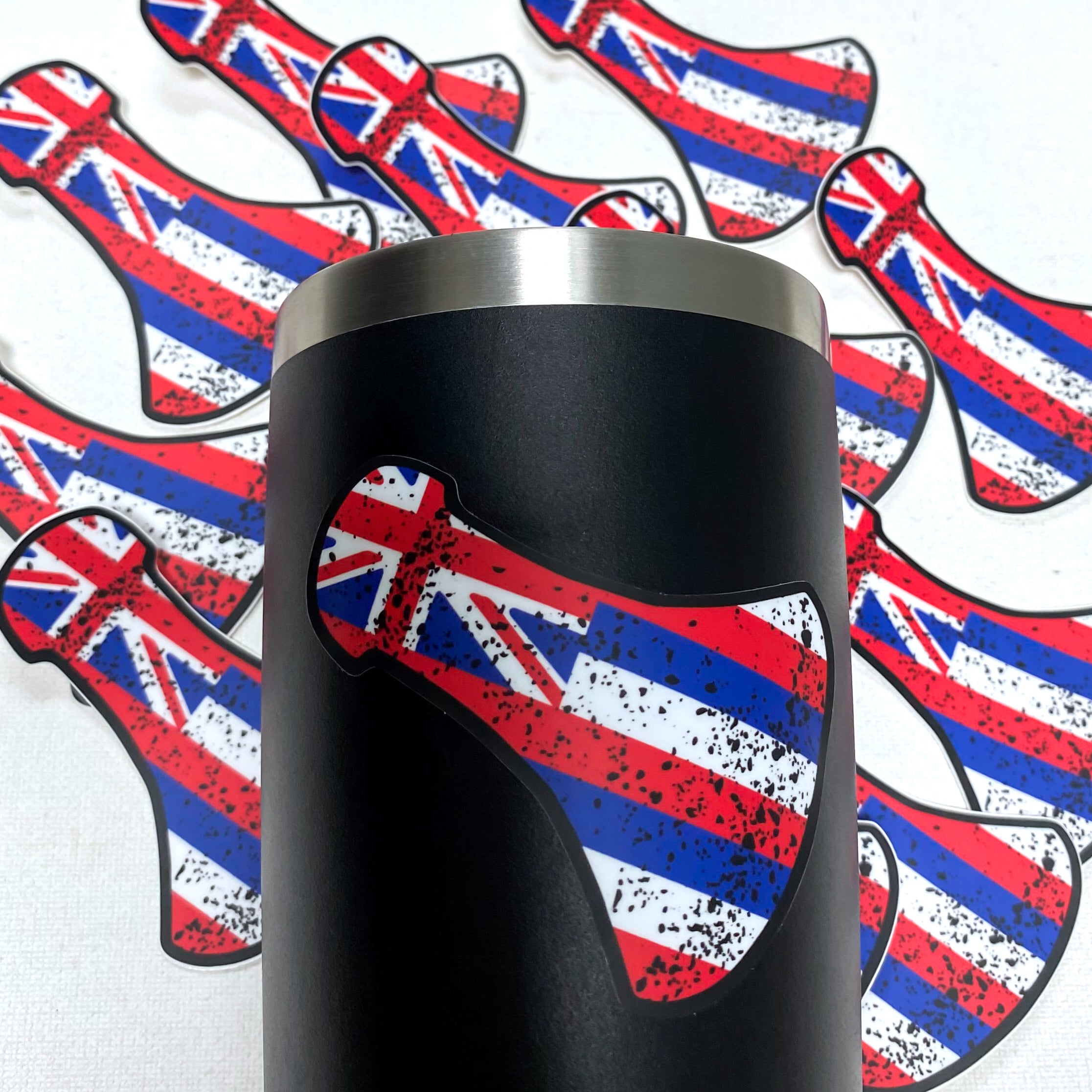 Close-up of a black tumbler decorated with a sticker shaped like a Hawaiian war club featuring the Hawaiian flag with a distressed black splatter pattern. In the background, multiple identical stickers are scattered on a white surface.
