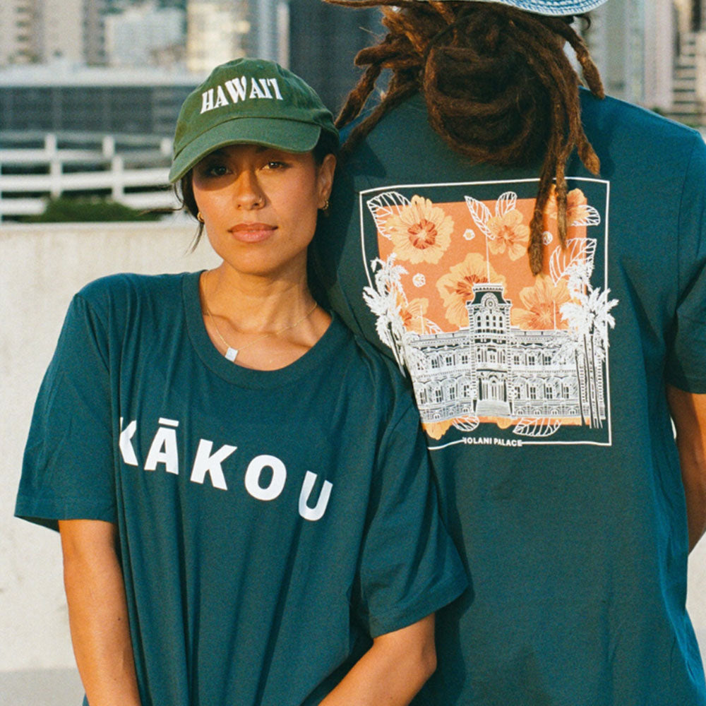 A woman wearing a dark teal t-shirt with the word "KĀKOU" in bold white letters stands beside a man. He wears a matching shirt featuring an illustration of ʻIolani Palace framed by tropical flowers and palm trees on an orange background. The woman also wears a green cap with "HAWAII" embroidered in white.