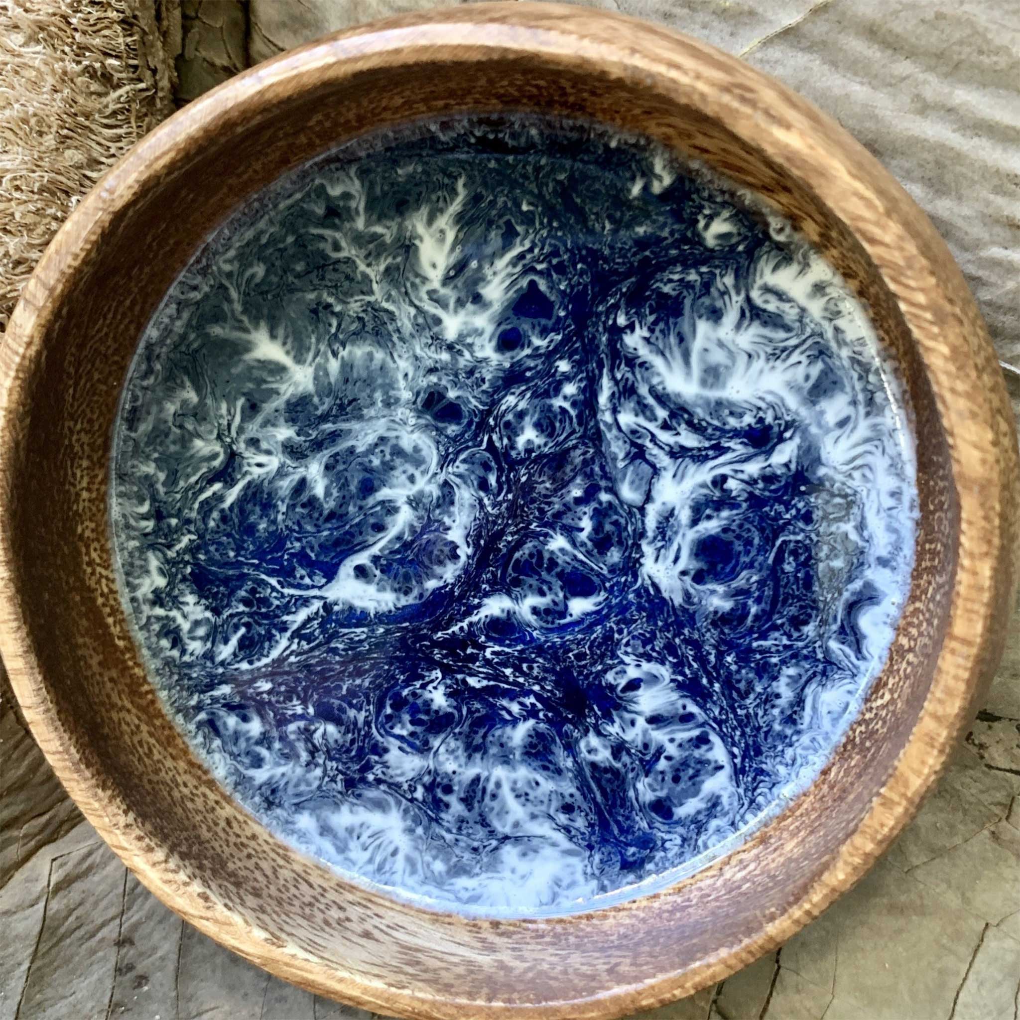 Top view of a wooden bowl featuring a blue and white resin design, evoking the look of turbulent ocean water, placed on a natural stone surface.