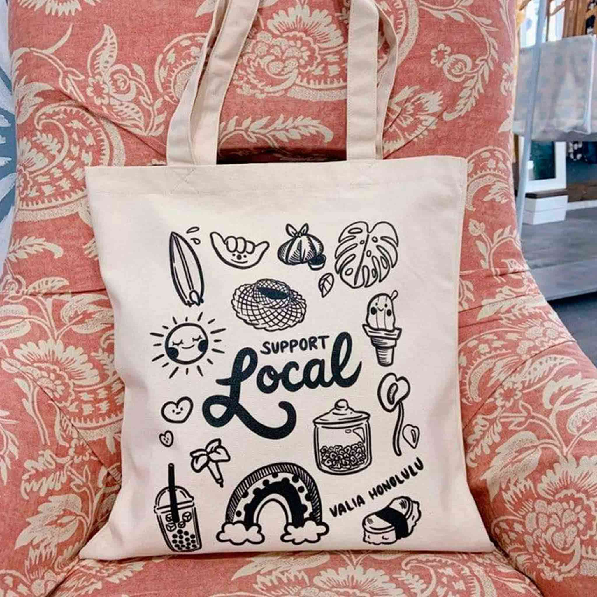 Canvas tote bag styled in a cozy boutique chair, showcasing its playful “Support Local” design filled with Hawai‘i-inspired doodles, from spam musubi to rainbow and shave ice. A charming, functional piece that radiates island pride and makes every errand feel like a celebration of community and Aloha.