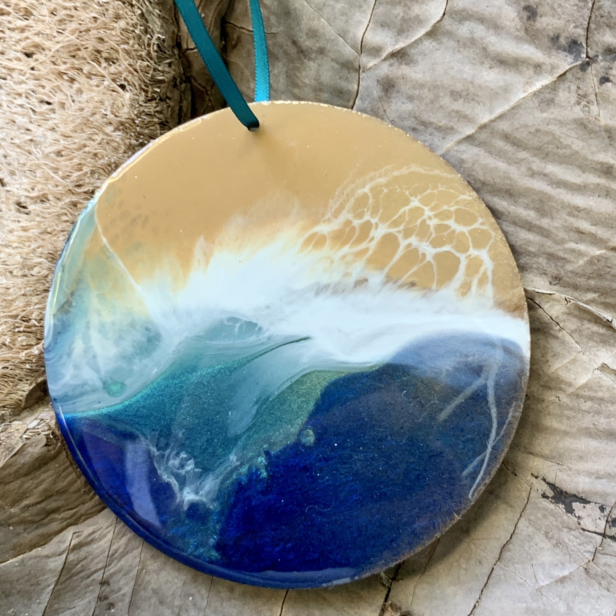 A handcrafted round resin ornament with a beach scene design. It features golden sand at the top, foamy white waves in the middle, and deep blue and green ocean tones below. The piece is glossy and hung with a teal ribbon, resting on natural textured paper and fiber.
