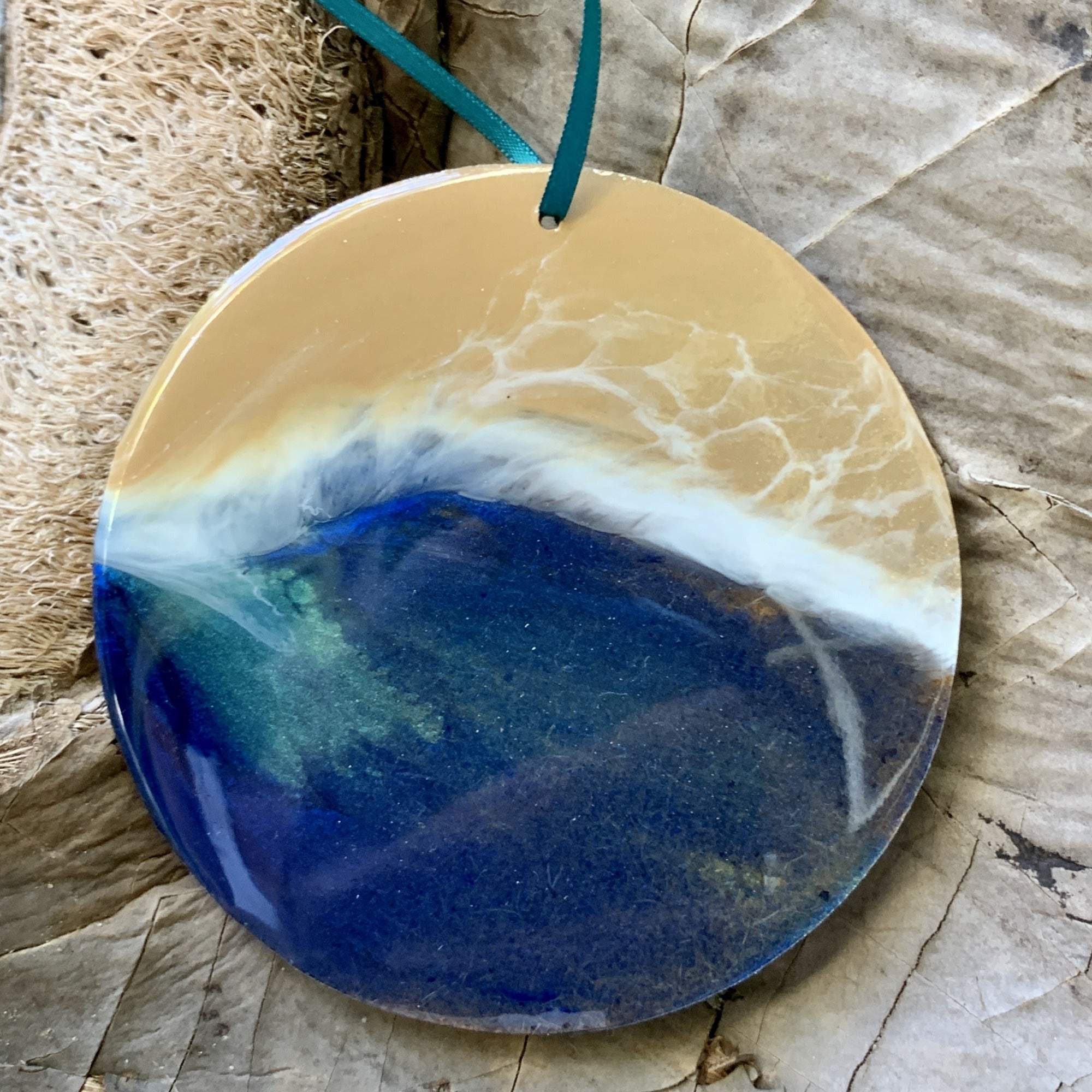 A circular ocean-inspired resin ornament with shimmering sand and layered ocean waves in blue, green, and white. The waves have a wispy, foamy texture, and the ornament hangs from a teal ribbon against a rustic background of dried palm bark.
