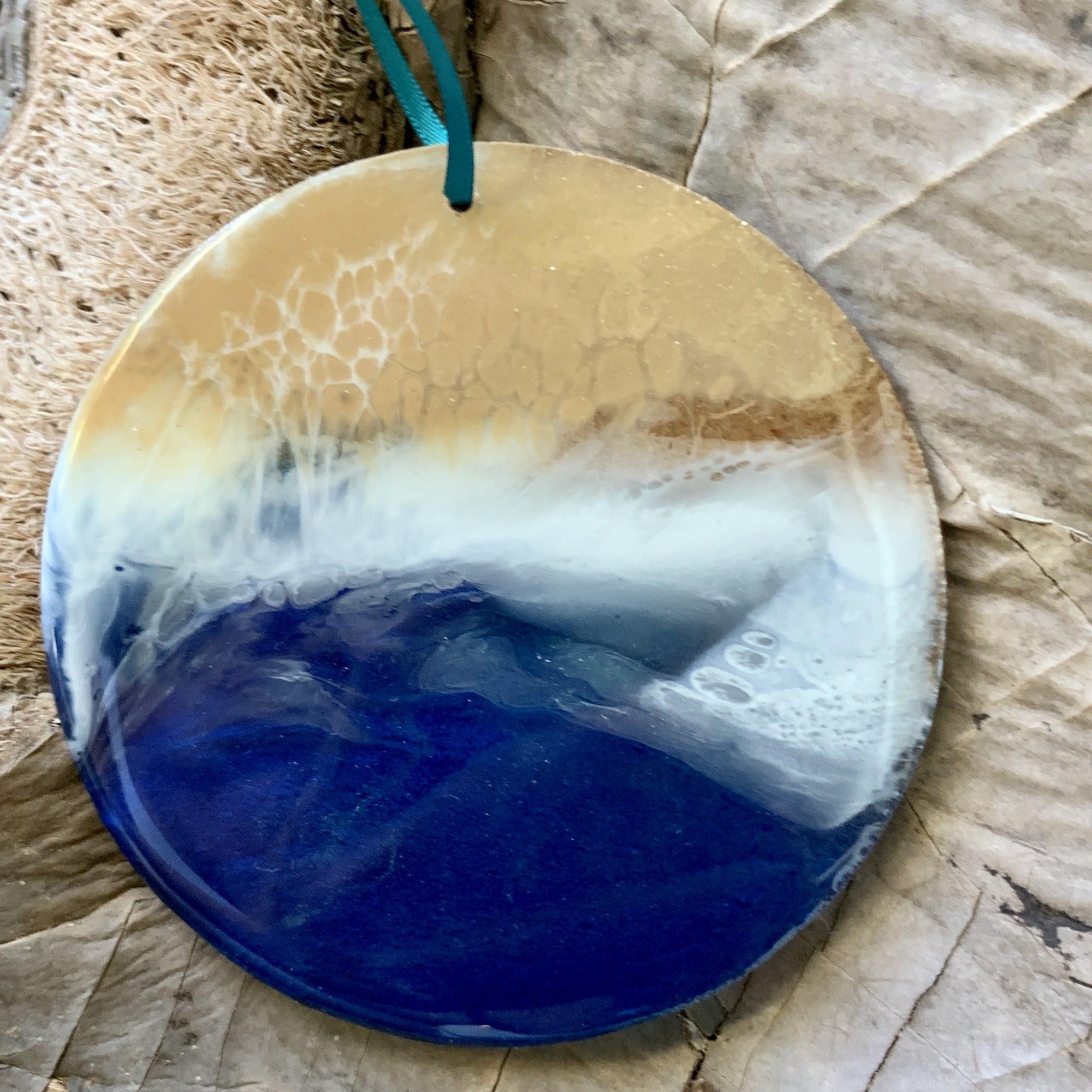 A handmade resin ornament with a realistic ocean wave effect, showing golden sand, white foam, and rich blue water. The top layer includes lacy wave patterns. A teal ribbon loops through the ornament, displayed on a background of brown crinkled natural fiber.