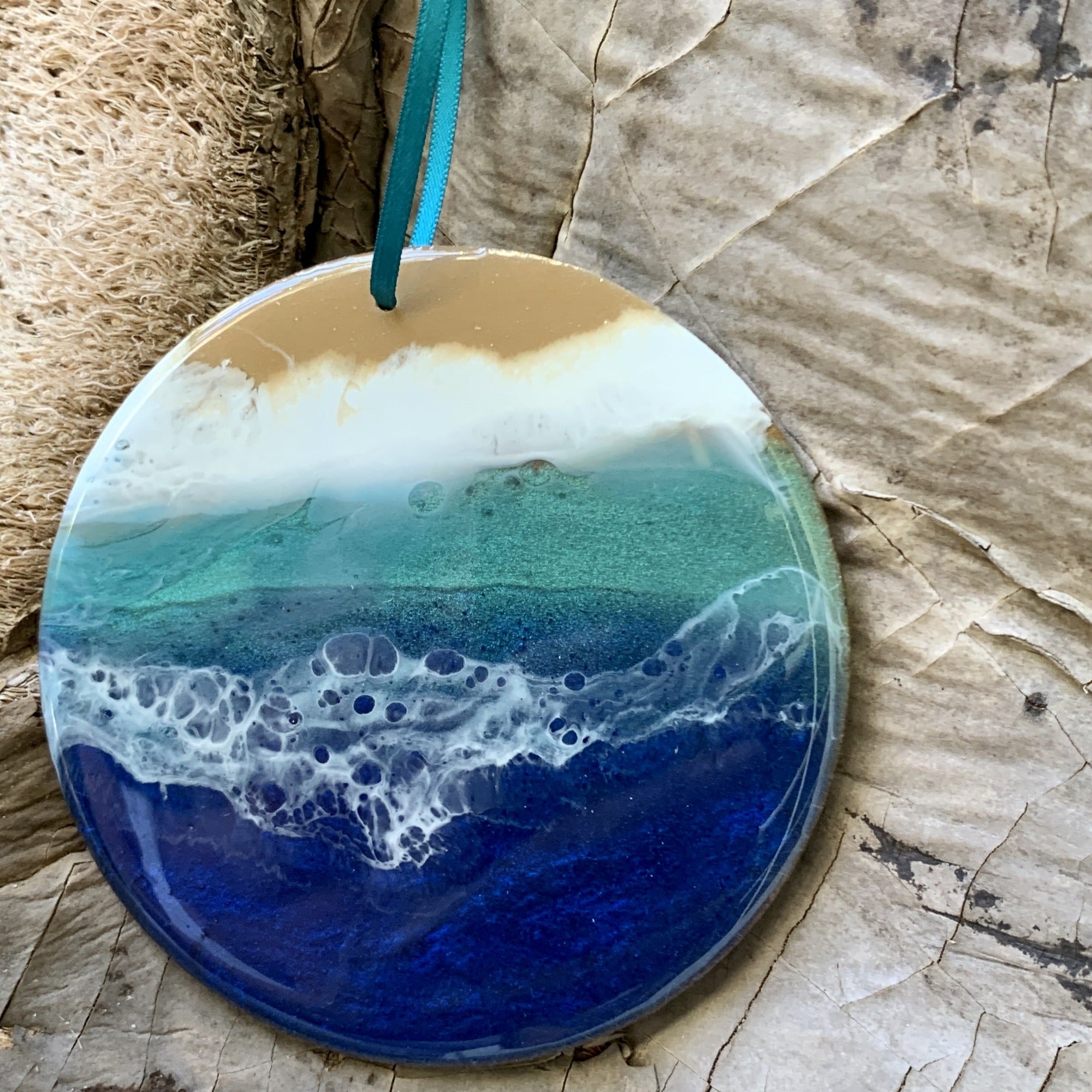 A round resin ornament featuring a vivid ocean scene with layered shades of deep blue, turquoise, and sandy beige, resembling waves crashing onto a shoreline. The ornament hangs from a teal ribbon and rests on a textured natural surface.