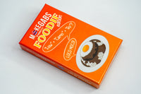 An orange rectangular product box for MEGABARS Foodie Edition, photographed on a plain light background. The box features large yellow and white 'FOODIE edition' text, a small Thai flag, and an illustration of a plate with a fried egg and rice.