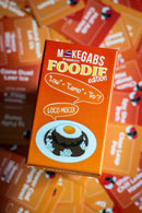 An orange product box for 'Mokegabs by Majuhen Est. 2020 Foodie edition' featuring a cartoon illustration of a chocolate pudding with a fried egg on a plate, surrounded by other colorful boxes in a shallow depth of field.