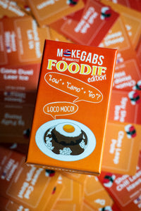 An orange product box for 'Mokegabs by Majuhen Est. 2020 Foodie edition' featuring a cartoon illustration of a chocolate pudding with a fried egg on a plate, surrounded by other colorful boxes in a shallow depth of field.
