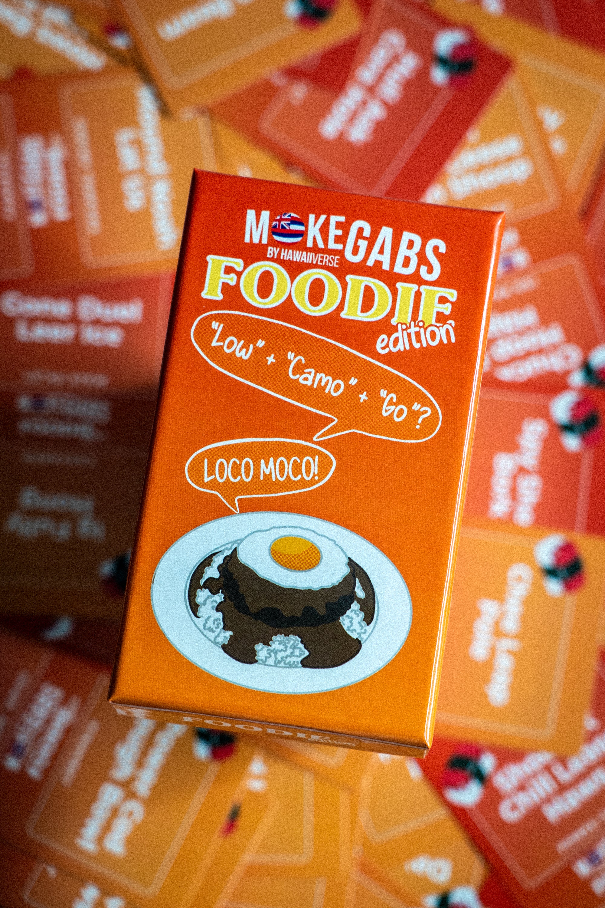 An orange product box for 'Mokegabs by Majuhen Est. 2020 Foodie edition' featuring a cartoon illustration of a chocolate pudding with a fried egg on a plate, surrounded by other colorful boxes in a shallow depth of field.