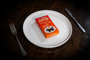 A small orange box of M&M's Foodie candy sits on a white plate, styled on a dark wooden table with a fork on the left and a knife on the right.