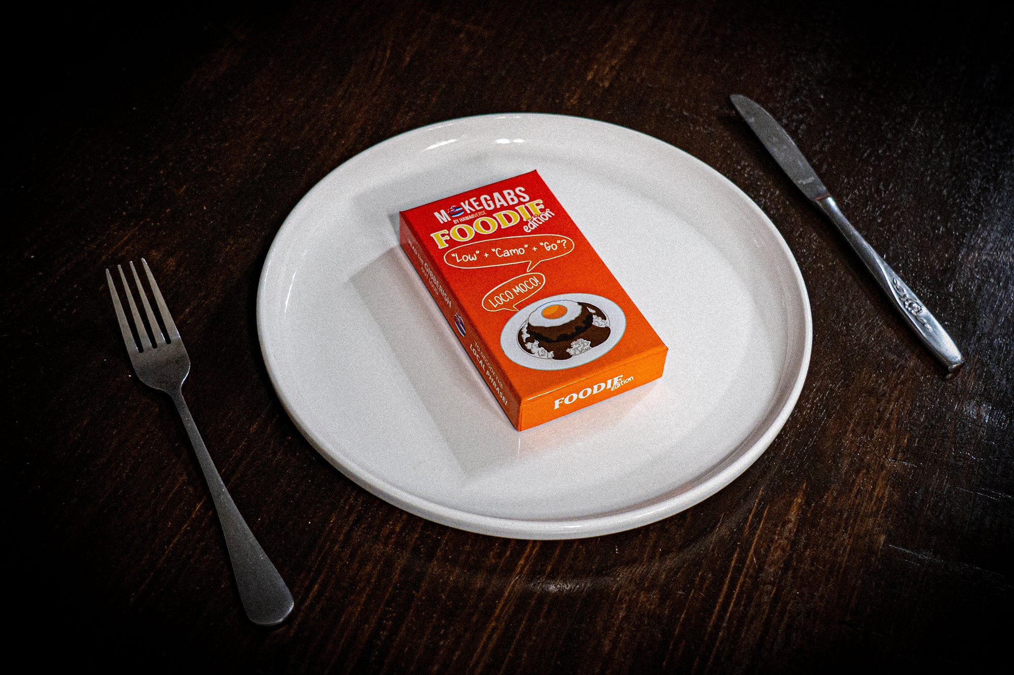 A small orange box of M&M's Foodie candy sits on a white plate, styled on a dark wooden table with a fork on the left and a knife on the right.