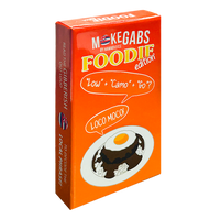 An orange rectangular product box for 'M-Kegabs by Hannah Reese Foodie edition' featuring a cartoon illustration of a chocolate cake with a fried egg on a plate and speech bubbles reading 'Low' + 'Cam' + 'Go'? and 'LOCO MOCA!'.
