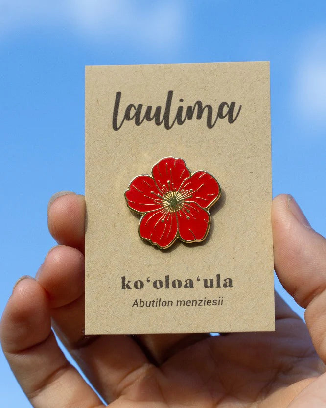 A hand holds a kraft-colored card with the brand name “laulima” at the top. Attached to the card is a red enamel pin shaped like a five-petal native Hawaiian flower. Below the pin, the card reads “ko‘oloa‘ula” and “Abutilon menziesii.” The sky is bright blue in the background.