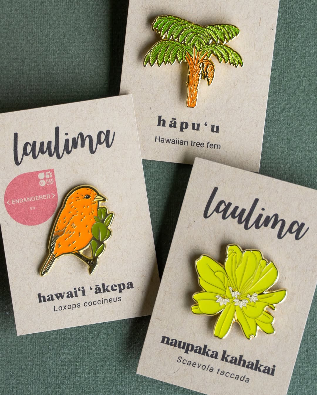 Three Hawaiian-themed enamel pins displayed on their cardboard backing cards against a dark green background. The collection features an orange Hawaiʻi ʻākepa bird pin labeled "laulima," a green and brown hāpuʻu Hawaiian tree fern pin, and a bright lime green naupaka kahakai flower pin also labeled "laulima."