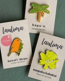 Three Hawaiian-themed enamel pins displayed on their cardboard backing cards against a dark green background. The collection features an orange Hawaiʻi ʻākepa bird pin labeled "laulima," a green and brown hāpuʻu Hawaiian tree fern pin, and a bright lime green naupaka kahakai flower pin also labeled "laulima."