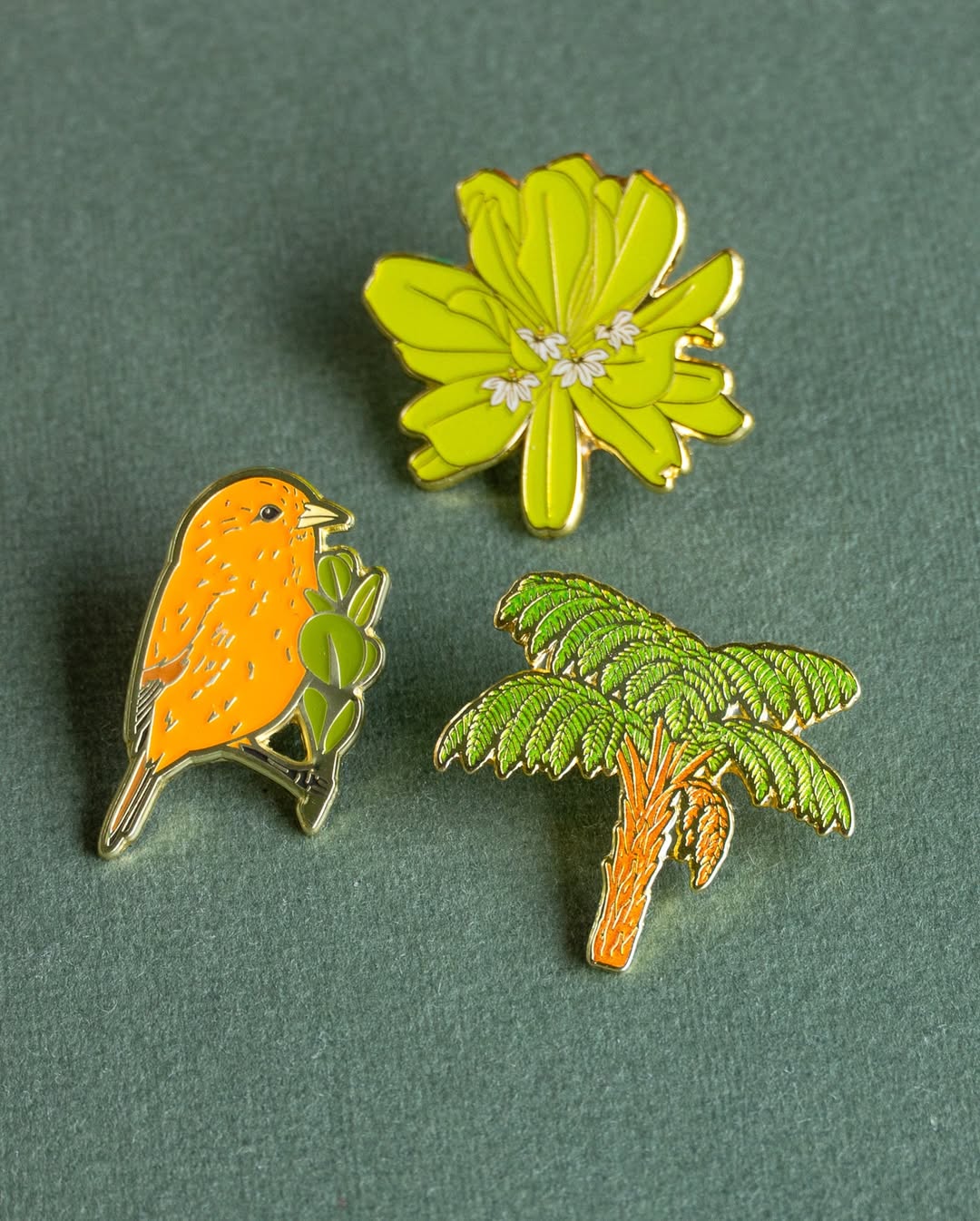 A top-down view of three gold-plated enamel pins arranged closely together on a textured green surface. The pins include an orange Hawaiʻi ʻākepa bird perched on a branch, a green hāpuʻu tree fern with an orange trunk, and a vibrant lime-green naupaka kahakai flower cluster.