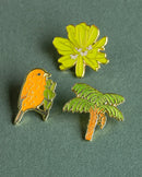 A top-down view of three gold-plated enamel pins arranged closely together on a textured green surface. The pins include an orange Hawaiʻi ʻākepa bird perched on a branch, a green hāpuʻu tree fern with an orange trunk, and a vibrant lime-green naupaka kahakai flower cluster.