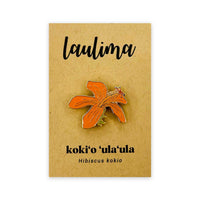 An orange and gold enamel pin shaped like a hibiscus flower, mounted on a brown card with the text 'laulima' and 'koki’o ʻula ʻula Hibiscus kokio' below it.